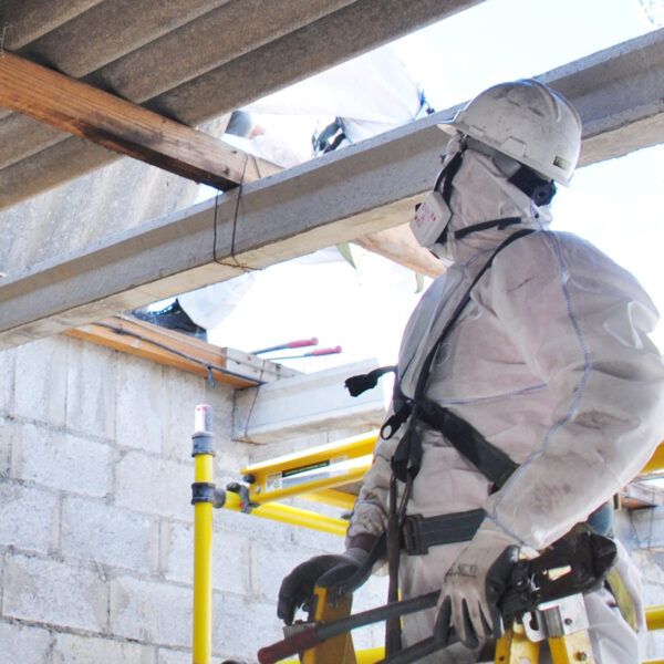 Worker wearing protective suit and respirator inspecting asbestos roof sheets from scaffolding.