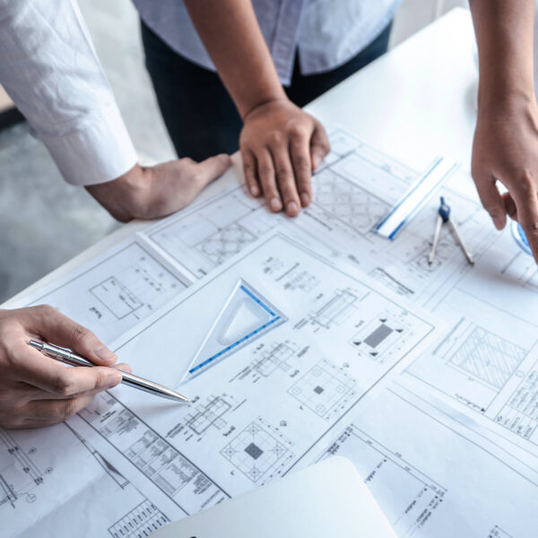 Hands of construction professionals reviewing architectural drawings and plans on a table with safety equipment nearby.