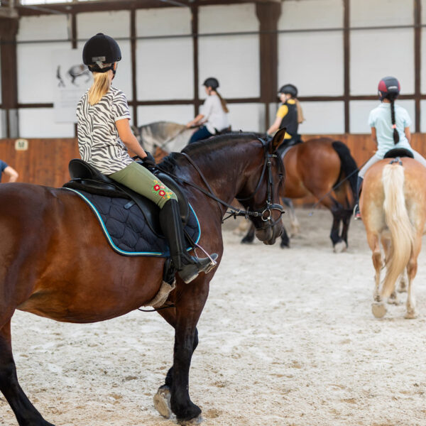 Group of horse riders wearing helmets training in an indoor equestrian arena.