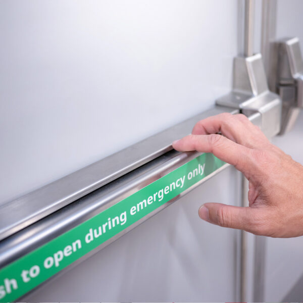 Person testing an emergency exit door push bar with a green “Push to open during emergency only” sign.