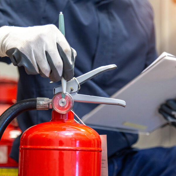 Person wearing gloves checking the pressure gauge on a red fire extinguisher while holding a clipboard.