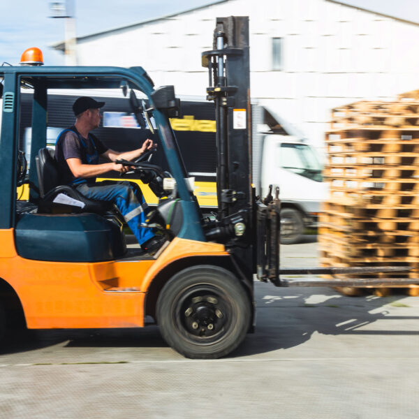 Worker driving a forklift carrying pallets in an industrial yard.