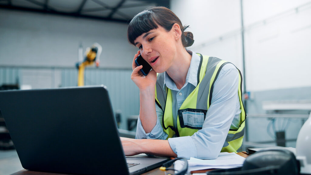 Warehouse supervisor wearing a high-vis vest talking on the phone and using a laptop.